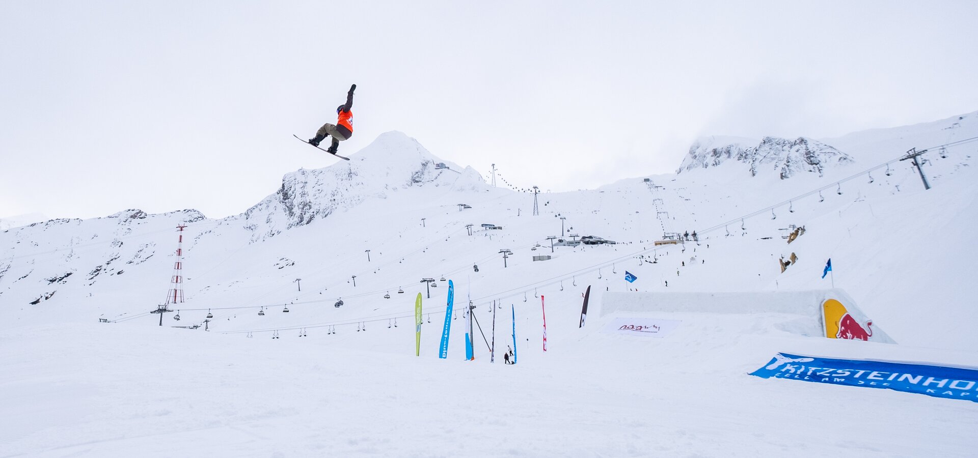 Snowboarderin springt vor verschneiter Bergkulisse über eine Rampe