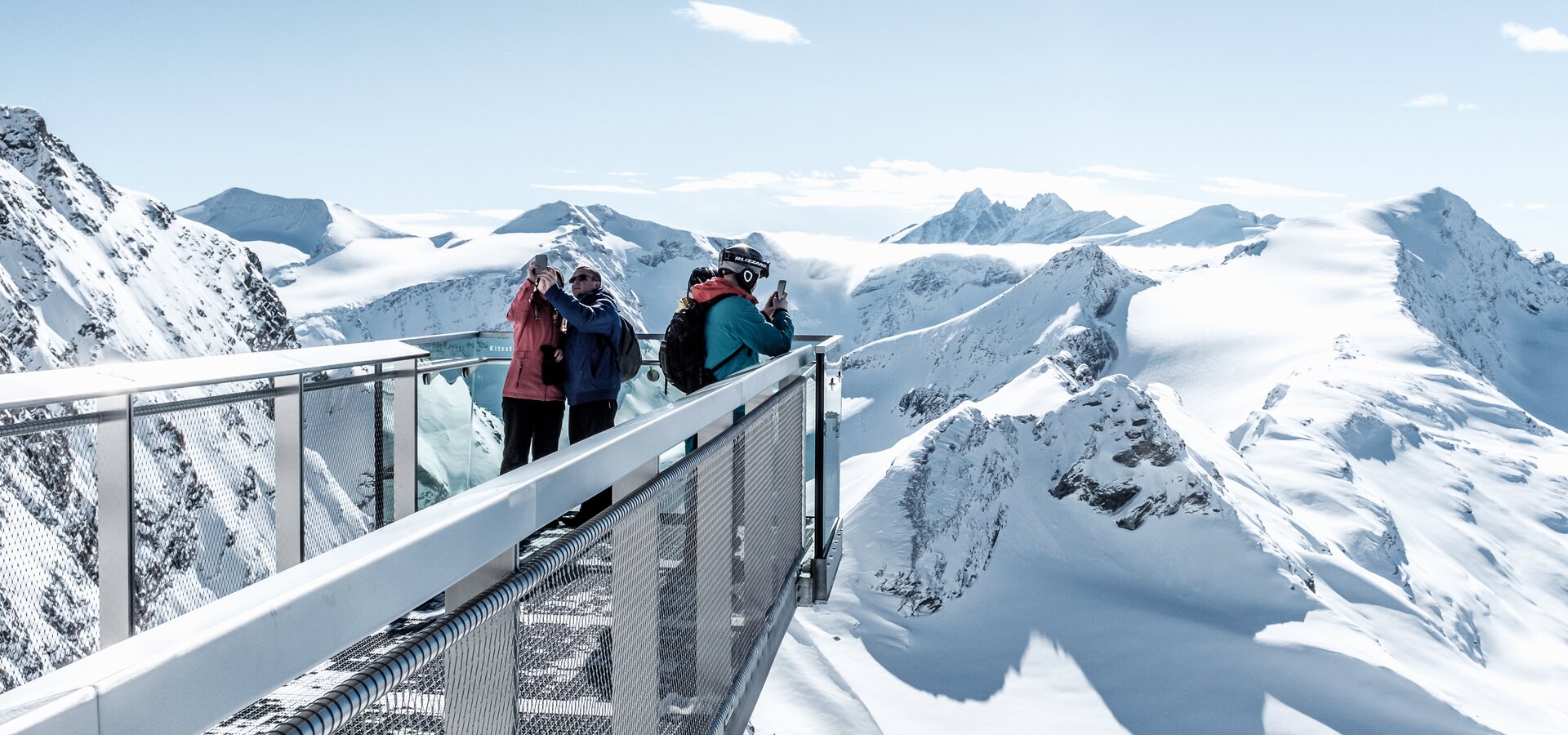 Menschen auf Aussichtsplattform in verschneiter Berglandschaft unter blauem Himmel