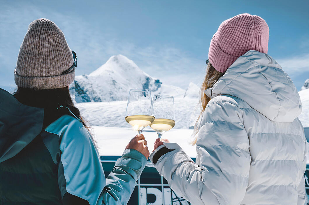 Two women clink wine glasses and admire the Kitzsteinhorn in a winter landscape | © Stefanie Oberhauser