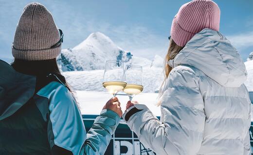 Zwei Damen stoßen mit Weingläsern an und betrachten das Kitzsteinhorn in winterlicher Landschaft | © Stefanie Oberhauser