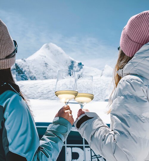 Zwei Damen stoßen mit Weingläsern an und betrachten das Kitzsteinhorn in winterlicher Landschaft | © Stefanie Oberhauser