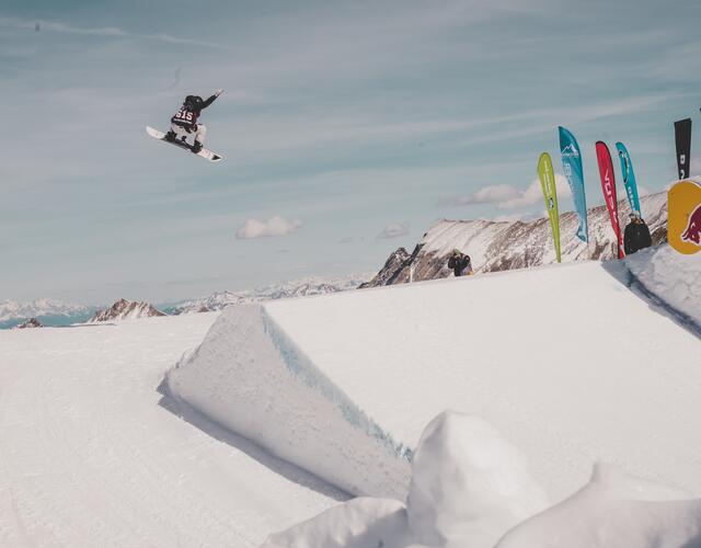 Snowboarder springt vor schneebedeckter Bergkulisse über Rampe | © Ania Grzelka