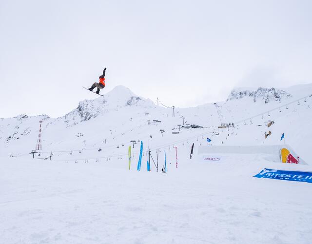 Snowboarderin springt vor verschneiter Bergkulisse über eine Rampe