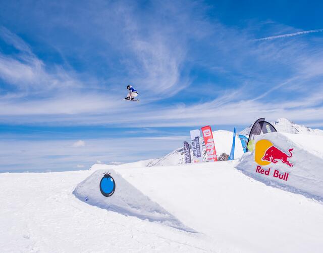 Snowboarderin springt über Schanze vor blauem Himmel und Red Bull Logo