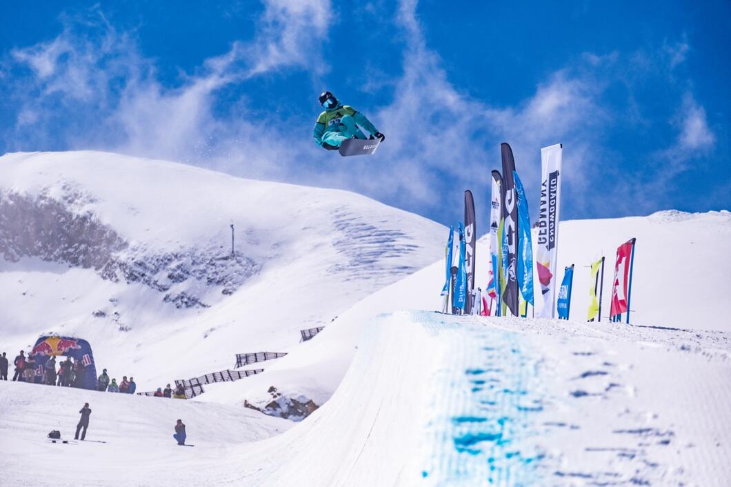 Snowboarder springt vor schneebedeckter Bergkulisse über eine Rampe