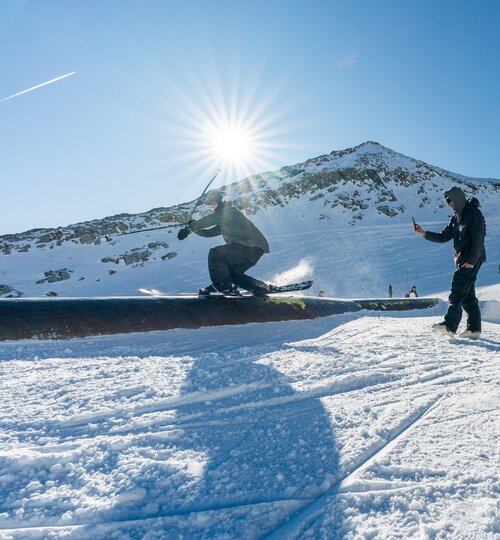 Ein Snowboarder führt im Snowpark am Kitzsteinhorn einen Trick aus, im Hintergrund filmt ihn jemand, neben einer Beach-Flag mit ‚Downdays Snowpark-Tour‘-Logo | © Stubai Photos by Klaus Polzer