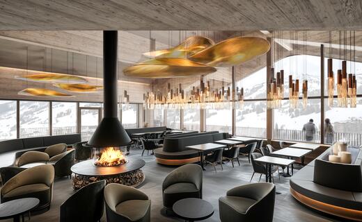 Innenansicht eines Restaurants mit Kamin, Tischen, Stühlen und goldfarbenen Lampen unter einer Holzdecke