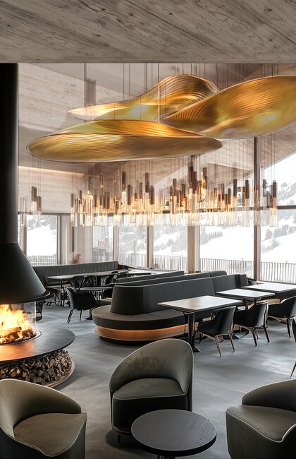 Innenansicht eines Restaurants mit Kamin, Tischen, Stühlen und goldfarbenen Lampen unter einer Holzdecke