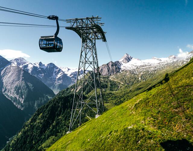 Seilbahn fährt über grüne Hänge, im Hintergrund schneebedeckte Berge unter blauem Himmel