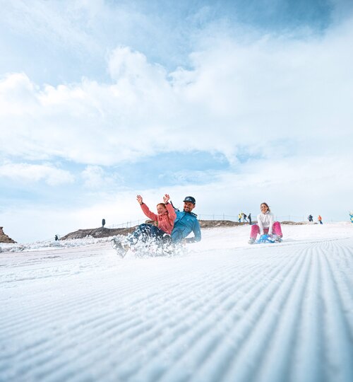Two people sledding on snow, another person sitting on a sled | © Kitzsteinhorn 
