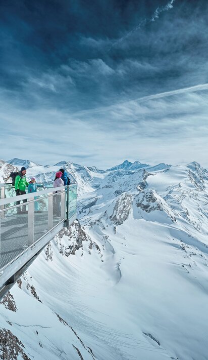 Personen auf Aussichtsplattform in verschneiter Berglandschaft unter bewölktem Himmel