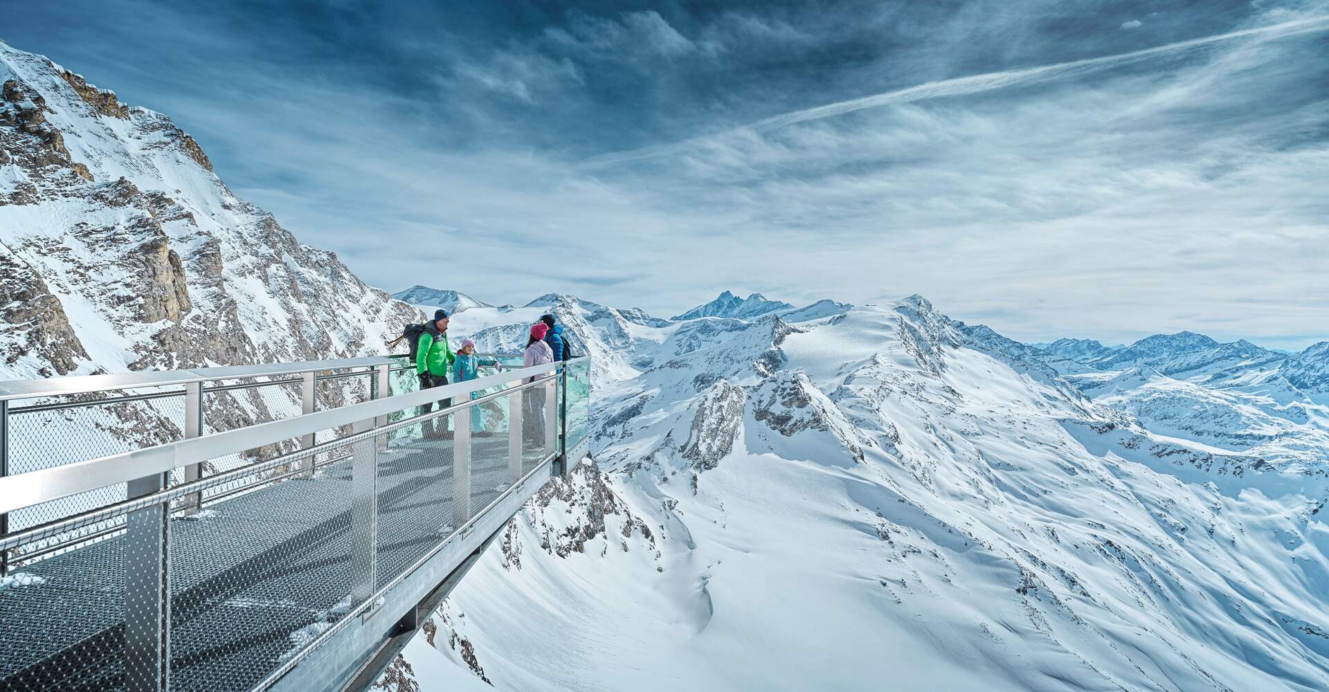 Personen auf Aussichtsplattform in verschneiter Berglandschaft unter bewölktem Himmel
