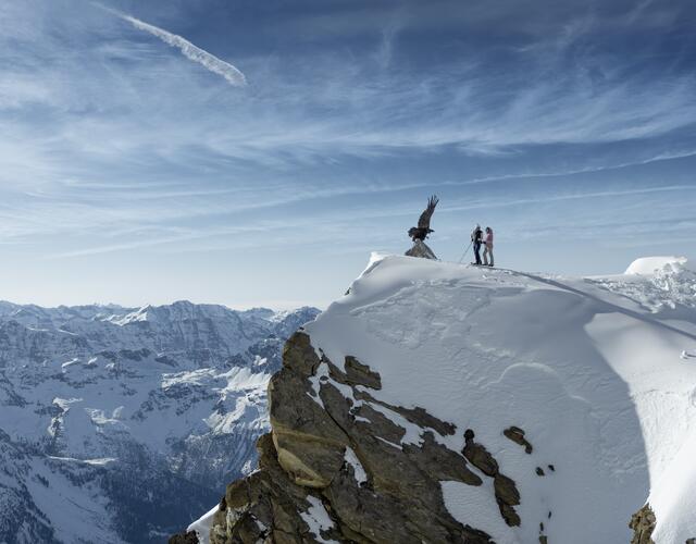A woman and a man in winter hiking gear stand at the viewpoint with a bronze eagle at the Kitzsteinhorn looking at the snowy Hohe Tauern National Park | © Kitzsteinhorn 