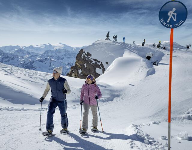 Winter landscape at the Kitzsteinhorn, a woman and a man with snowshoes walk a winter trail, in the background other people at a viewpoint enjoy the snowy scenery | © Kitzsteinhorn 