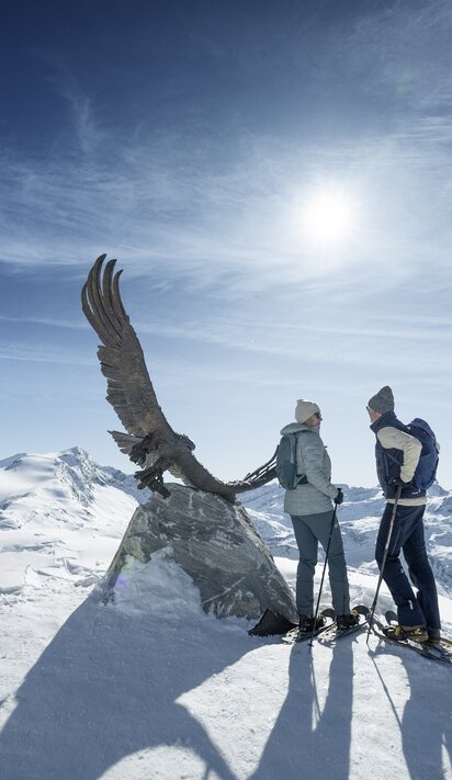 Eine Dame und ein Herr in Winterwanderkleidung stehen am Aussichtspunkt mit bronzenem Adler am Kitzsteinhorn und blicken auf den winterlichen Nationalpark Hohe Tauern