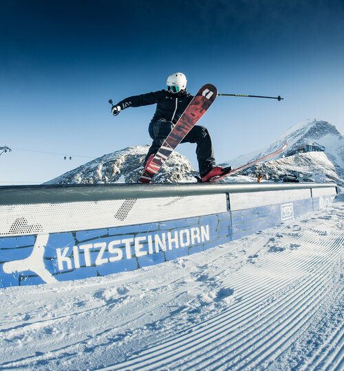 Skifahrer springt auf Kitzsteinhorn-Geländer vor schneebedeckten Bergen